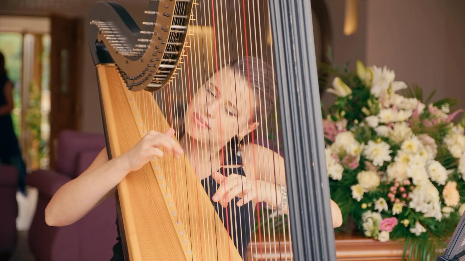 A young girl plays a wooden harp with a white and brown striped cord, her hands on the strings, in a room with a floral arrangement and a purple couch
