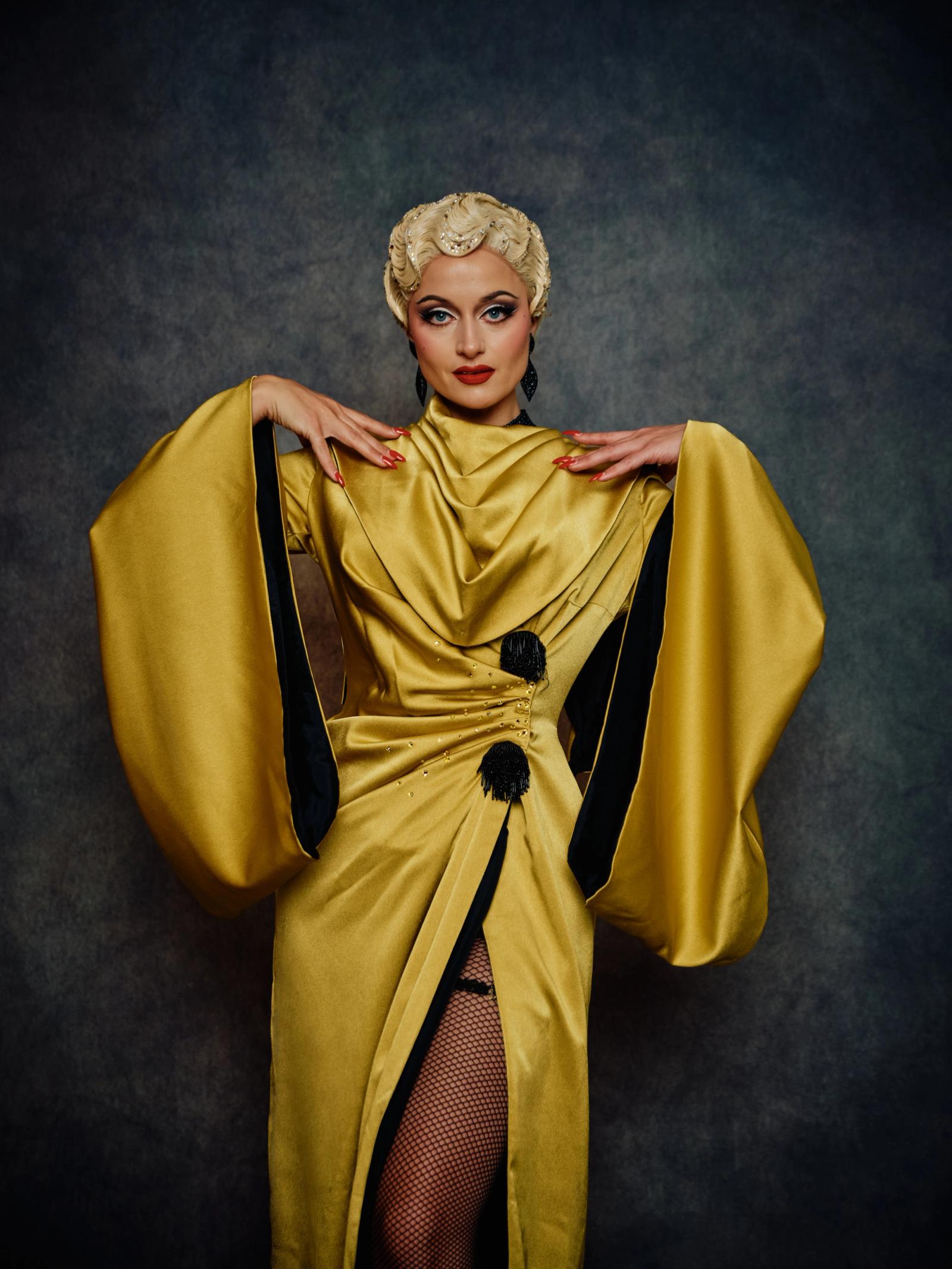A woman with blonde hair, wearing a vibrant yellow dress with black accents, stands confidently against a dark gray background. Her blonde hair cascad