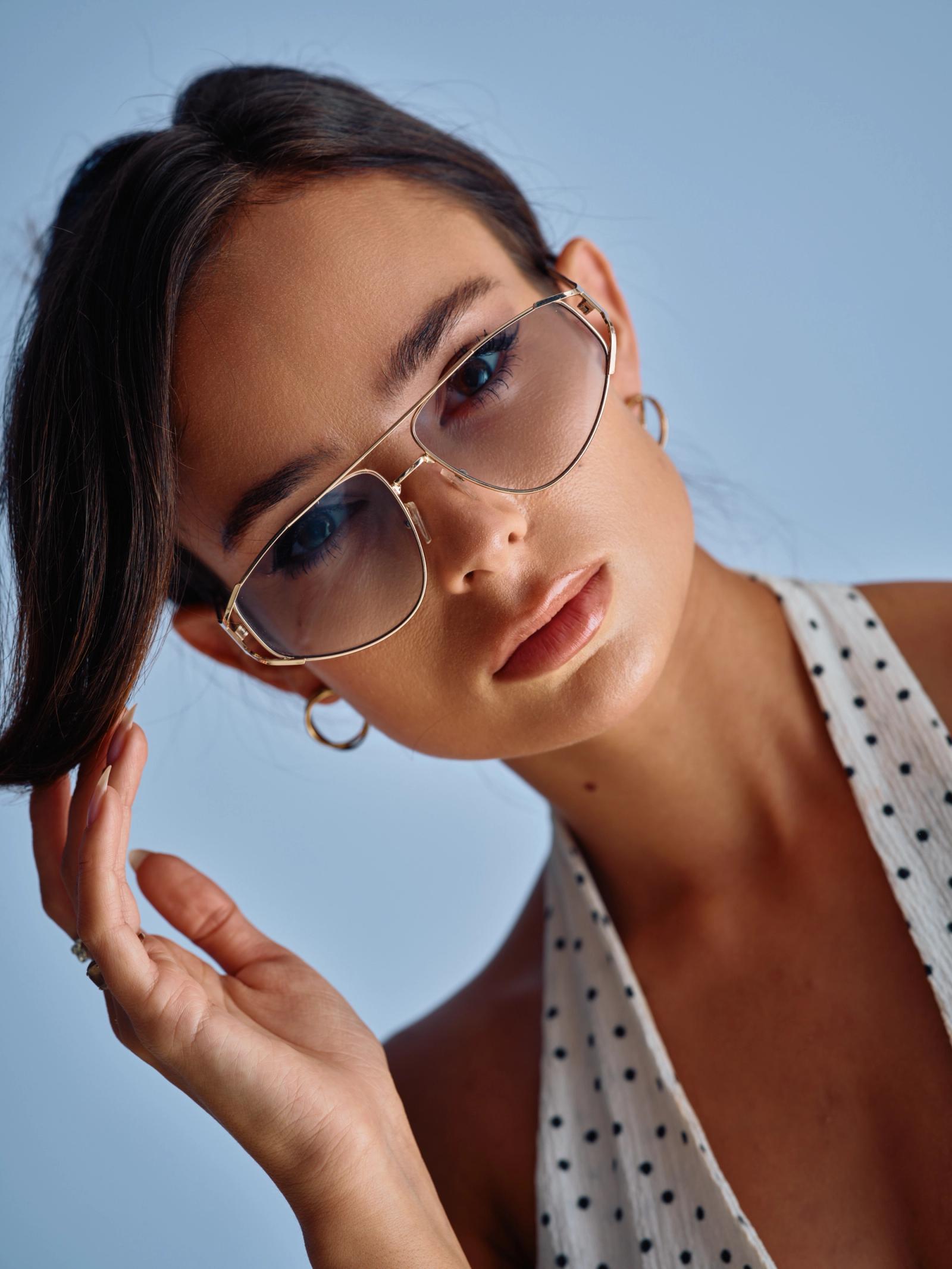 A young woman with long, dark hair and blue eyes wears gold-framed sunglasses and polka dot top, posing for a portrait against a light blue background
