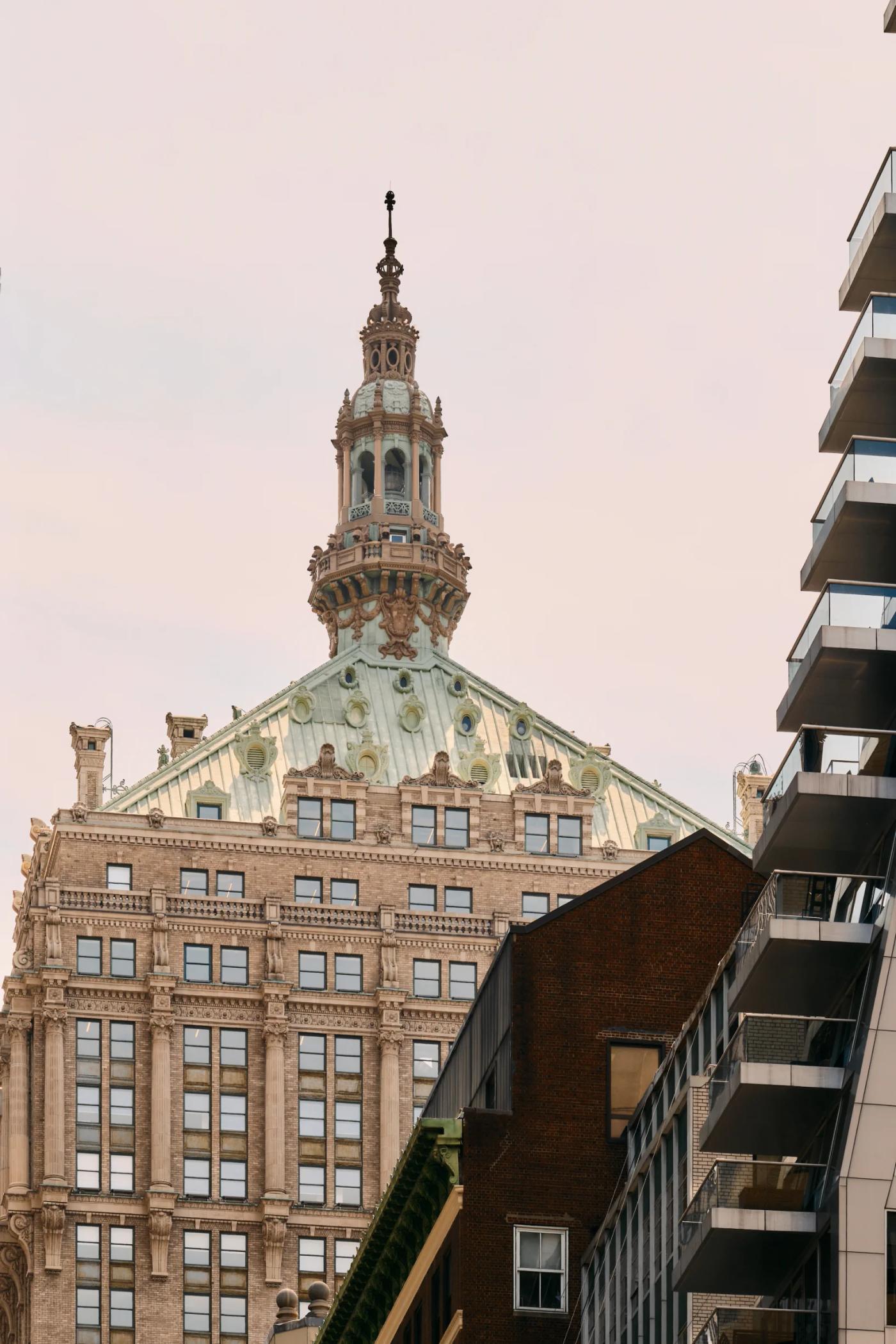 A majestic, ornate building with a green dome and white spire stands tall in a bustling cityscape, surrounded by modern buildings.
