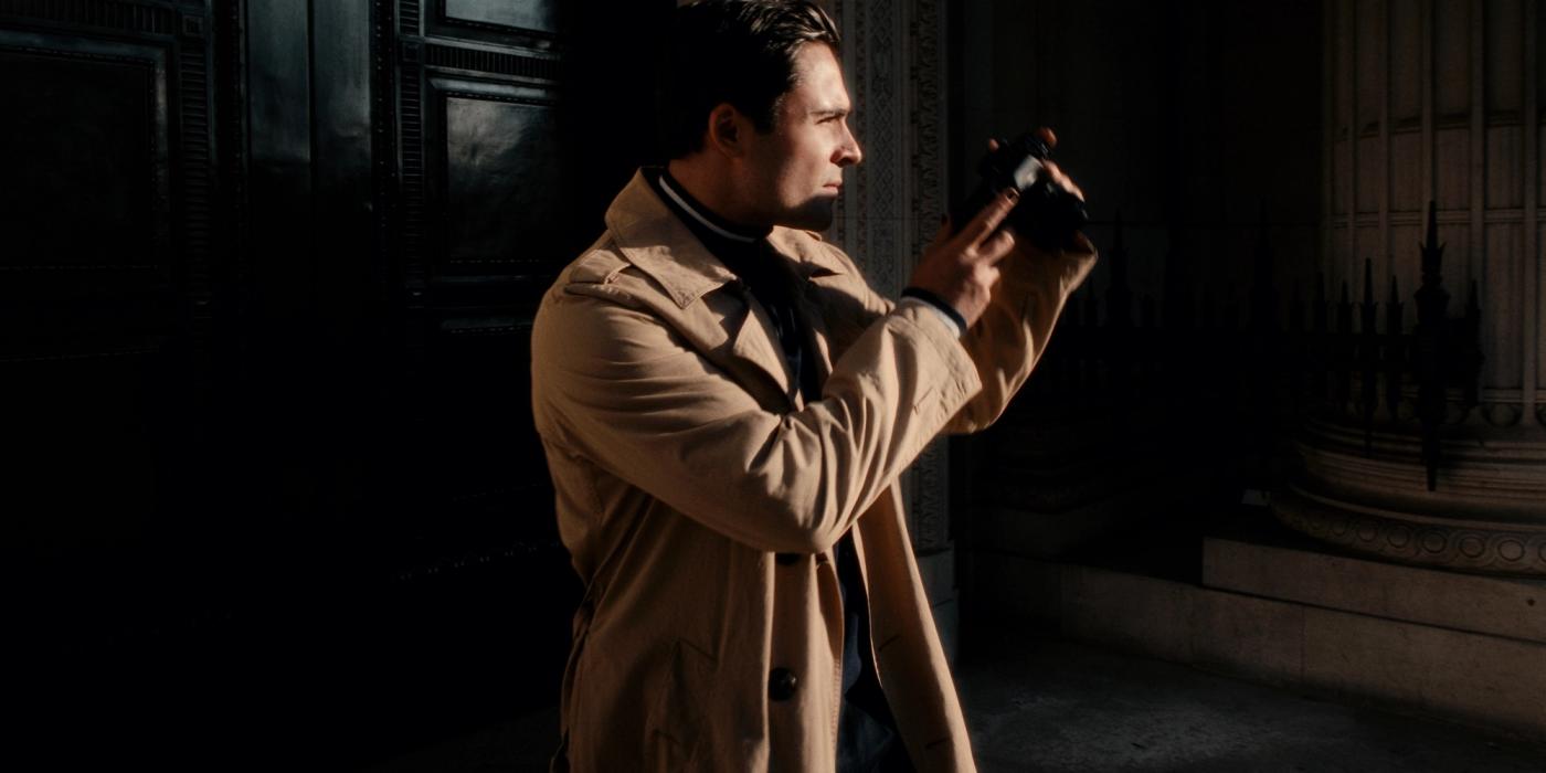 A man in a beige trench coat stands in front of a black door, holding a camera in his right hand and a camera in his left.