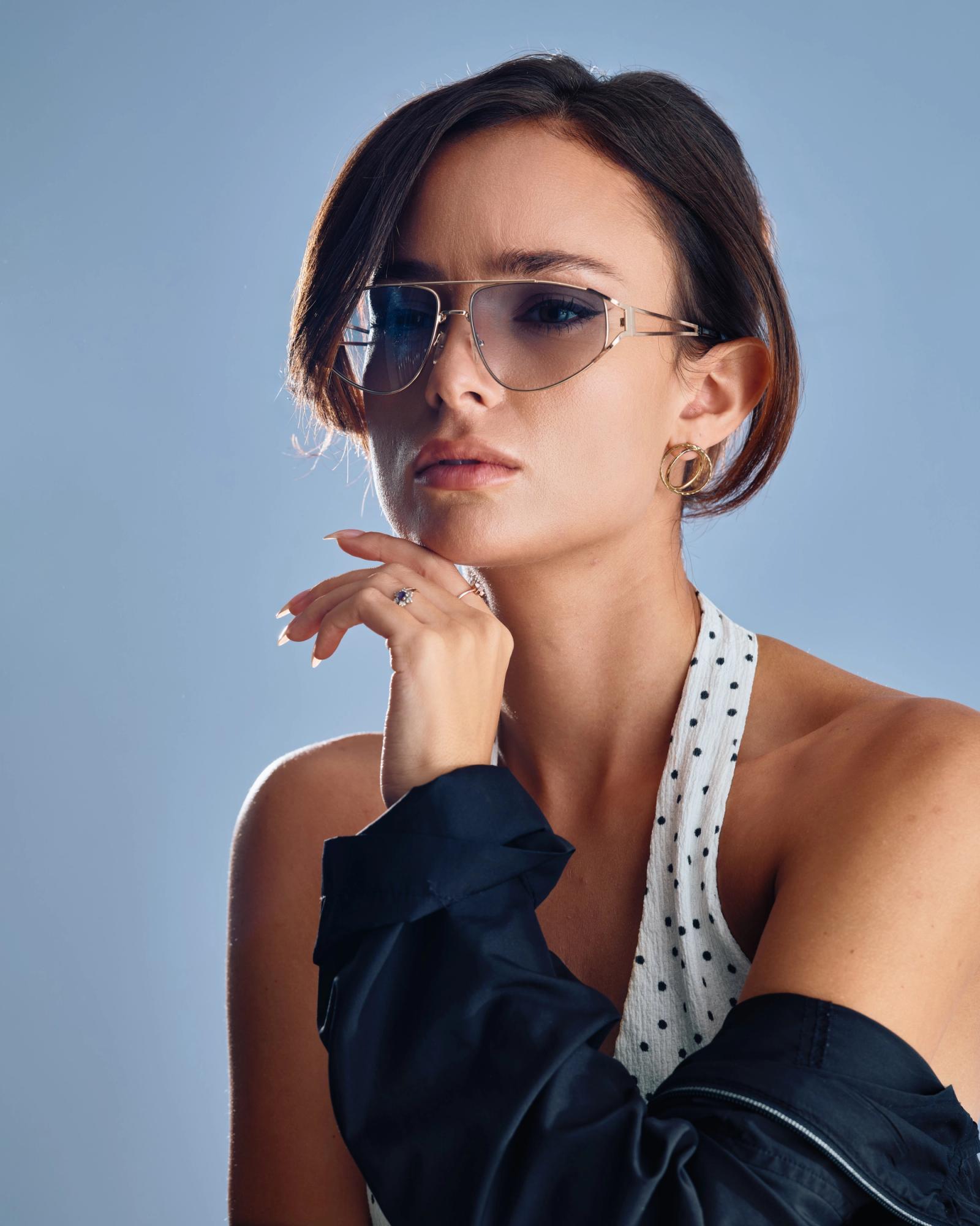A stylish woman with dark hair, wearing sunglasses, a white polka dot top, and a black jacket, poses elegantly with her hand on her chin and earrings.