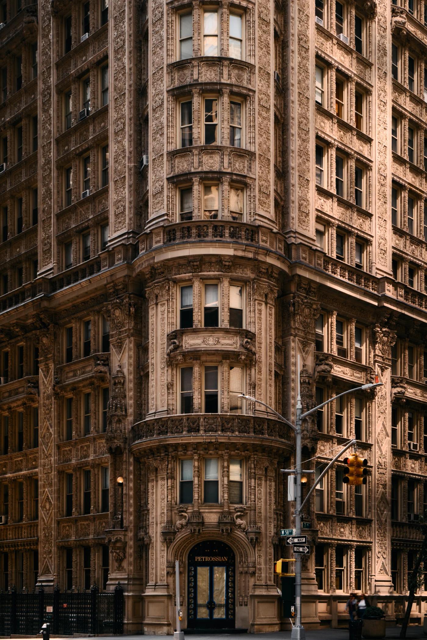 A large, ornate building with multiple levels and intricate details stands on a city street corner, surrounded by pedestrians and cars.
