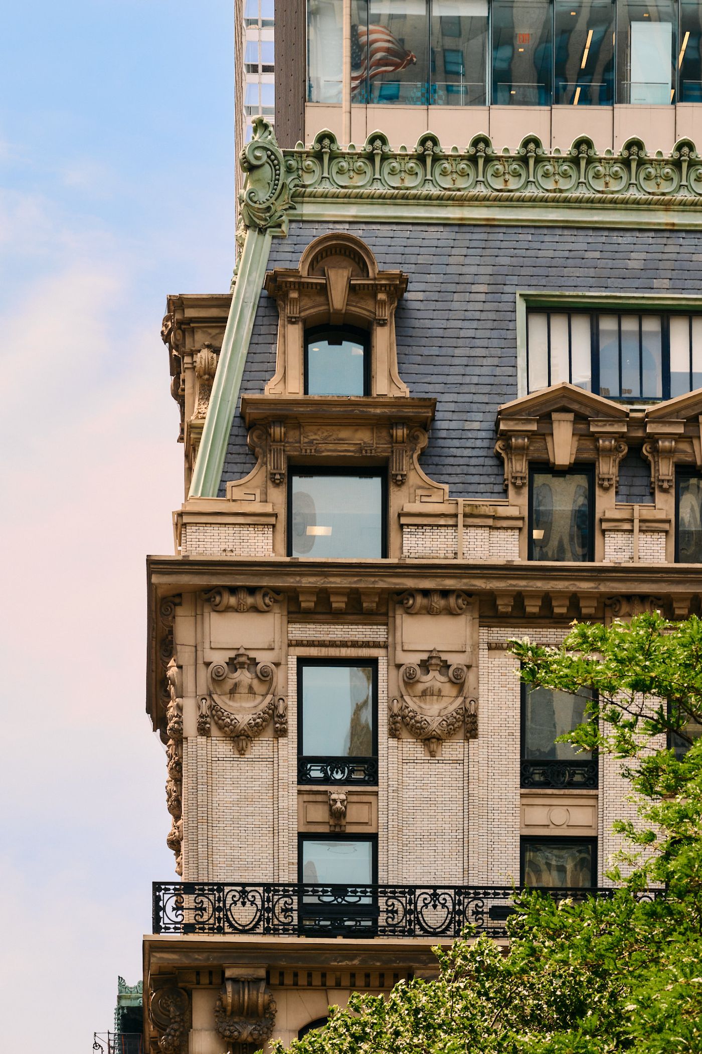 A beige stone building with ornate decorations, a black roof, and a balcony with a wrought iron railing stands tall against a pale blue sky.