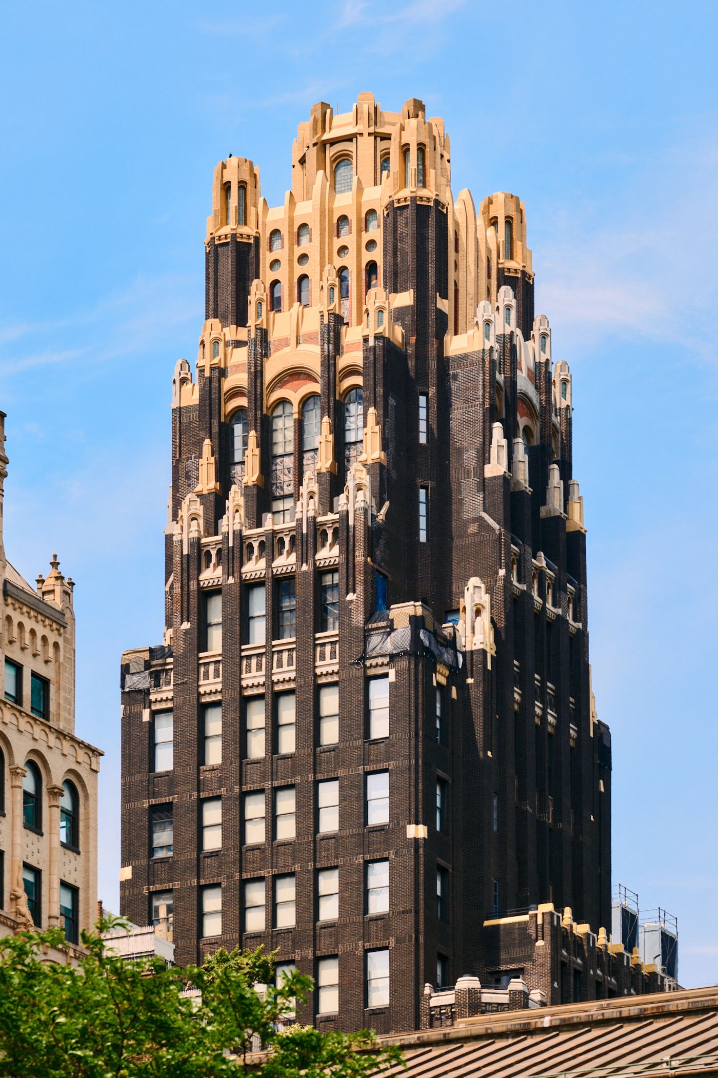 A tall, dark brown building with ornate spires and a golden-topped spire stands in a bustling city, surrounded by other buildings and trees.