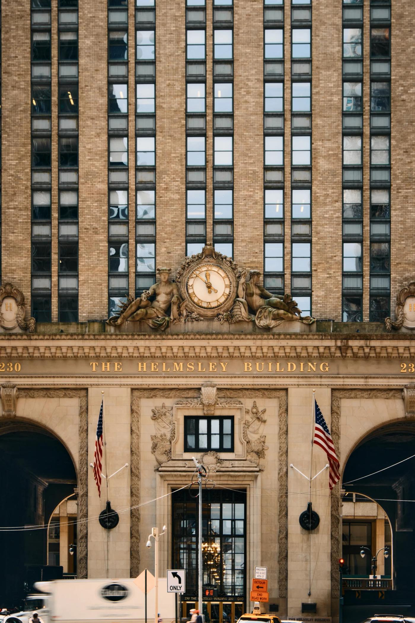 The image shows the entrance of the New York Stock Exchange, a large, historic building with a clock and American flags. People are walking around, an