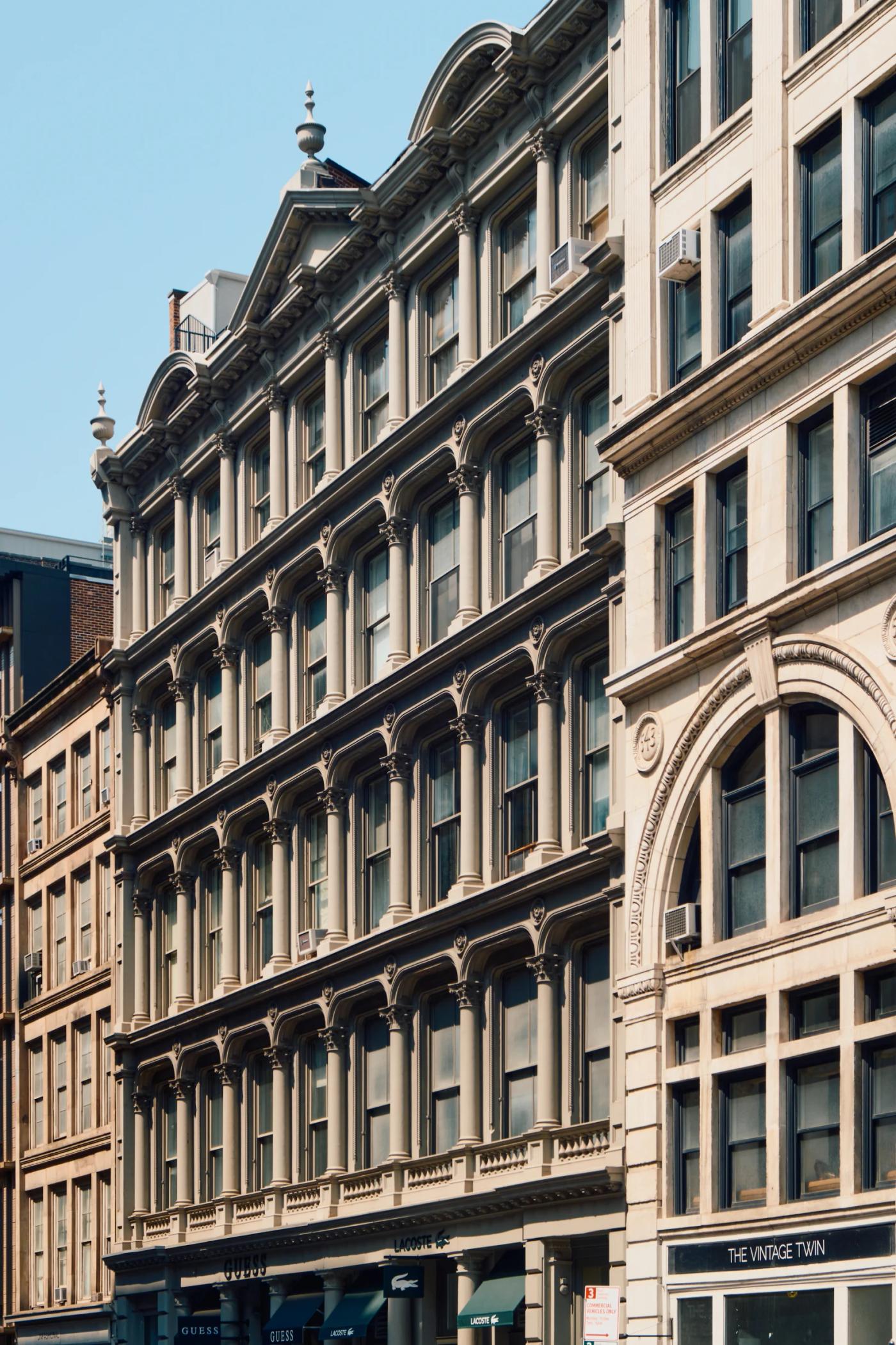 A bustling city street features a beige, ornate building with arched windows and a decorative pediment, surrounded by other buildings and shops.