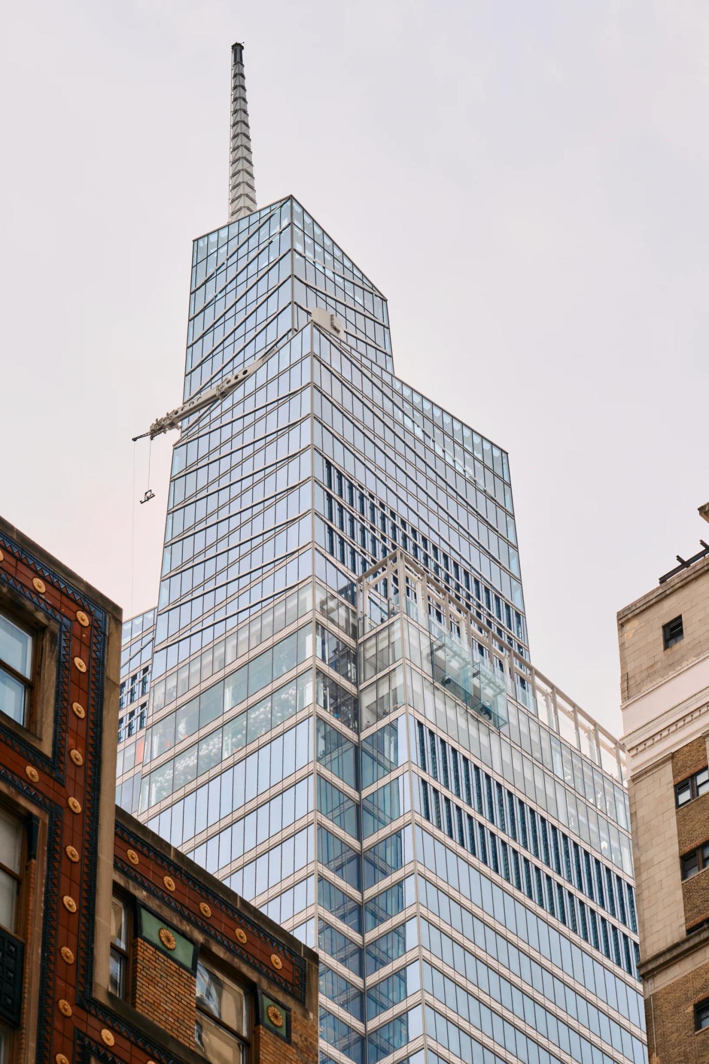 A skyscraper with a blue glass facade and a pointed spire dominates the cityscape, surrounded by other buildings and a clear sky.
