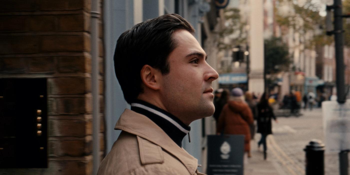 Actor Tom Hiddleston stands on a city street, wearing a beige trench coat and a black scarf, gazing off to the side. The street is filled with people 