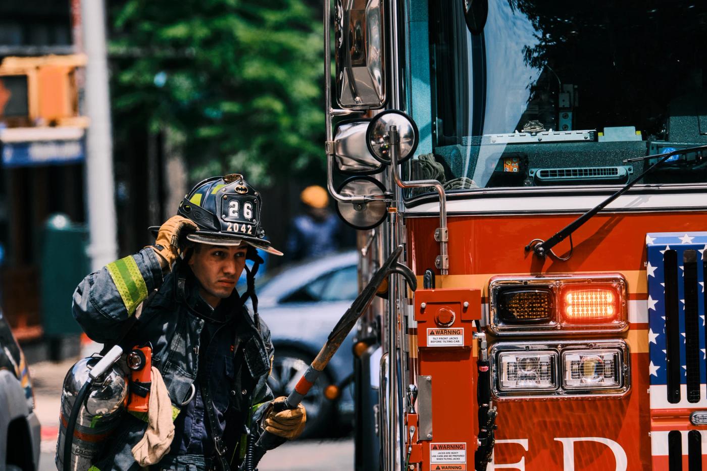 A firefighter in a green jacket and black cap, holding a fire hose, walks past a red fire truck with the number "10" on the side.