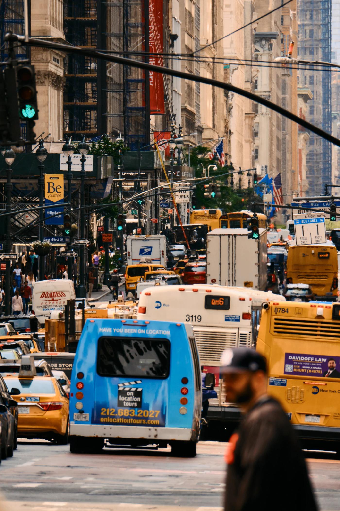 A bustling city street filled with various vehicles, including buses, taxis, and a blue and white bus, surrounded by tall buildings and a man walking 