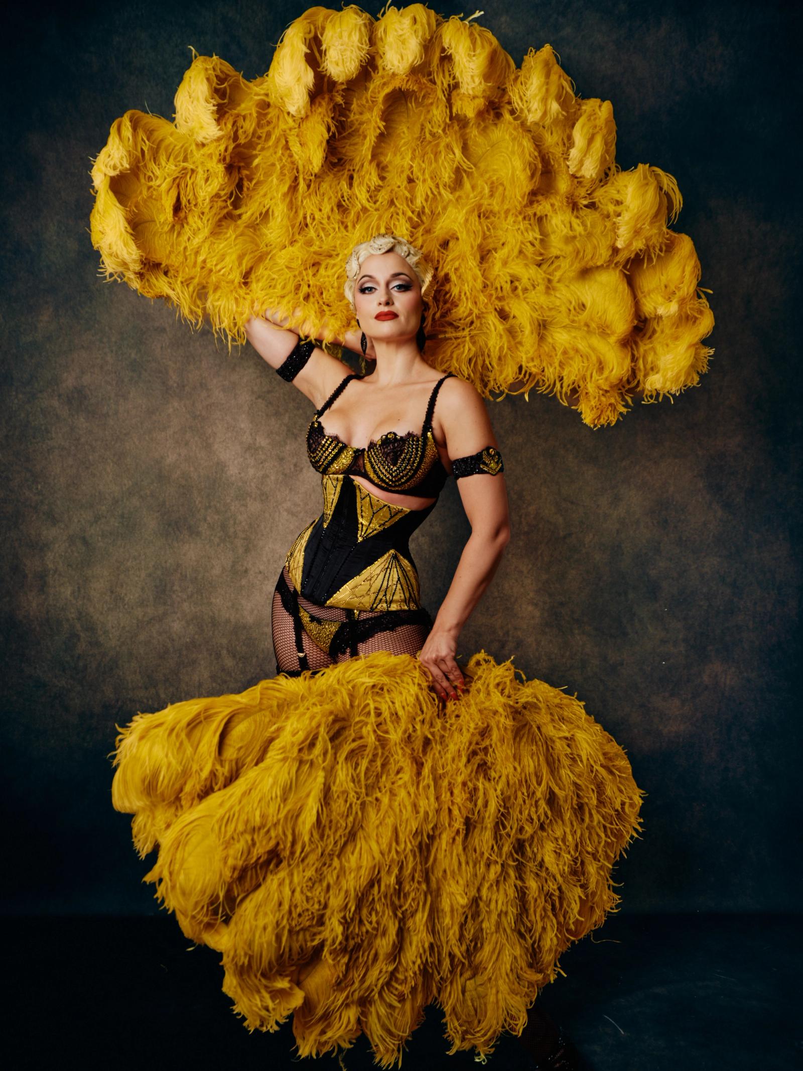 A woman in a black and gold costume, holding a large, feathered, and fluffed yellow feather fan, stands against a dark background.
