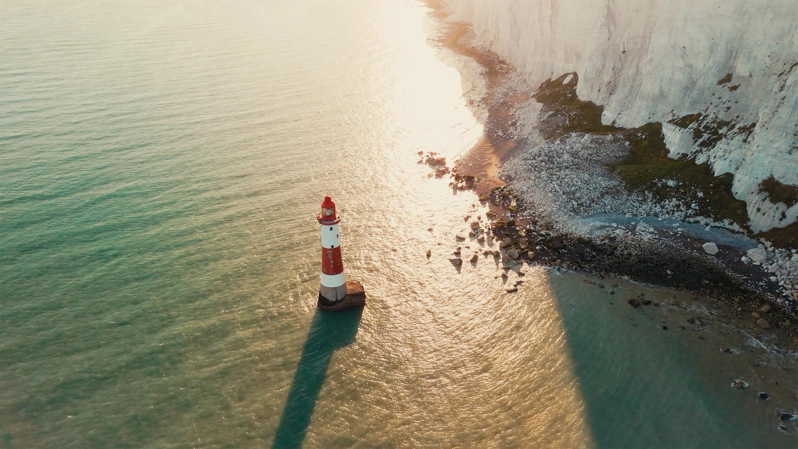 An aerial view of the Red and White Lighthouse on the cliffs of the English coast, with the sun casting a warm glow on the water and the cliff's white