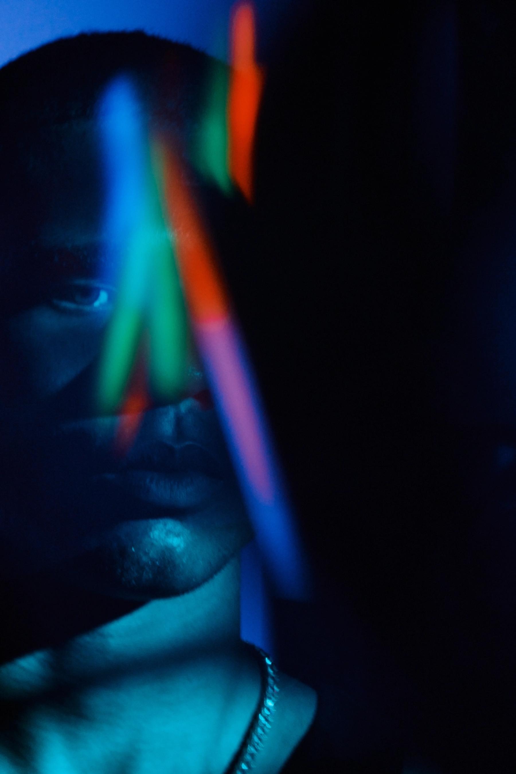 A man with a necklace and a bracelet is in a dimly lit room, with a colorful light reflection on his face.