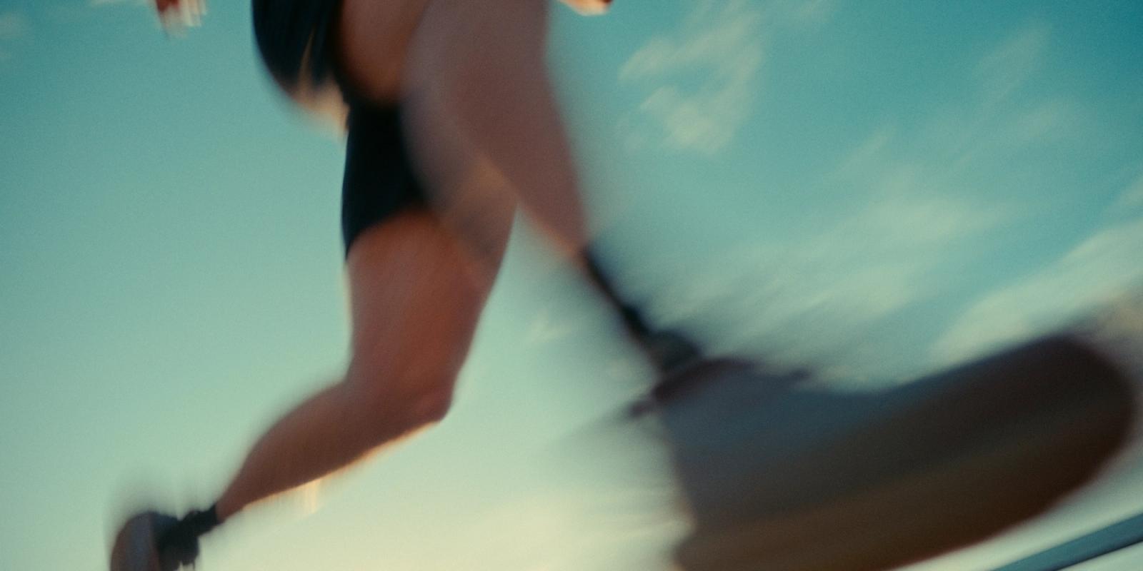 A blurred runner in black shorts and sneakers is mid-air, with legs extended and feet on a skateboard. The background is a serene blue sky with wispy 
