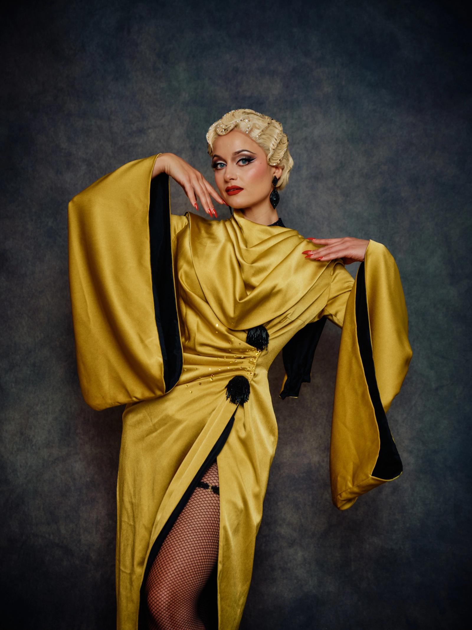 A woman in a golden robe with black accents, wearing black tights and fishnet stockings, poses confidently with her hands on her hips and her hair sty