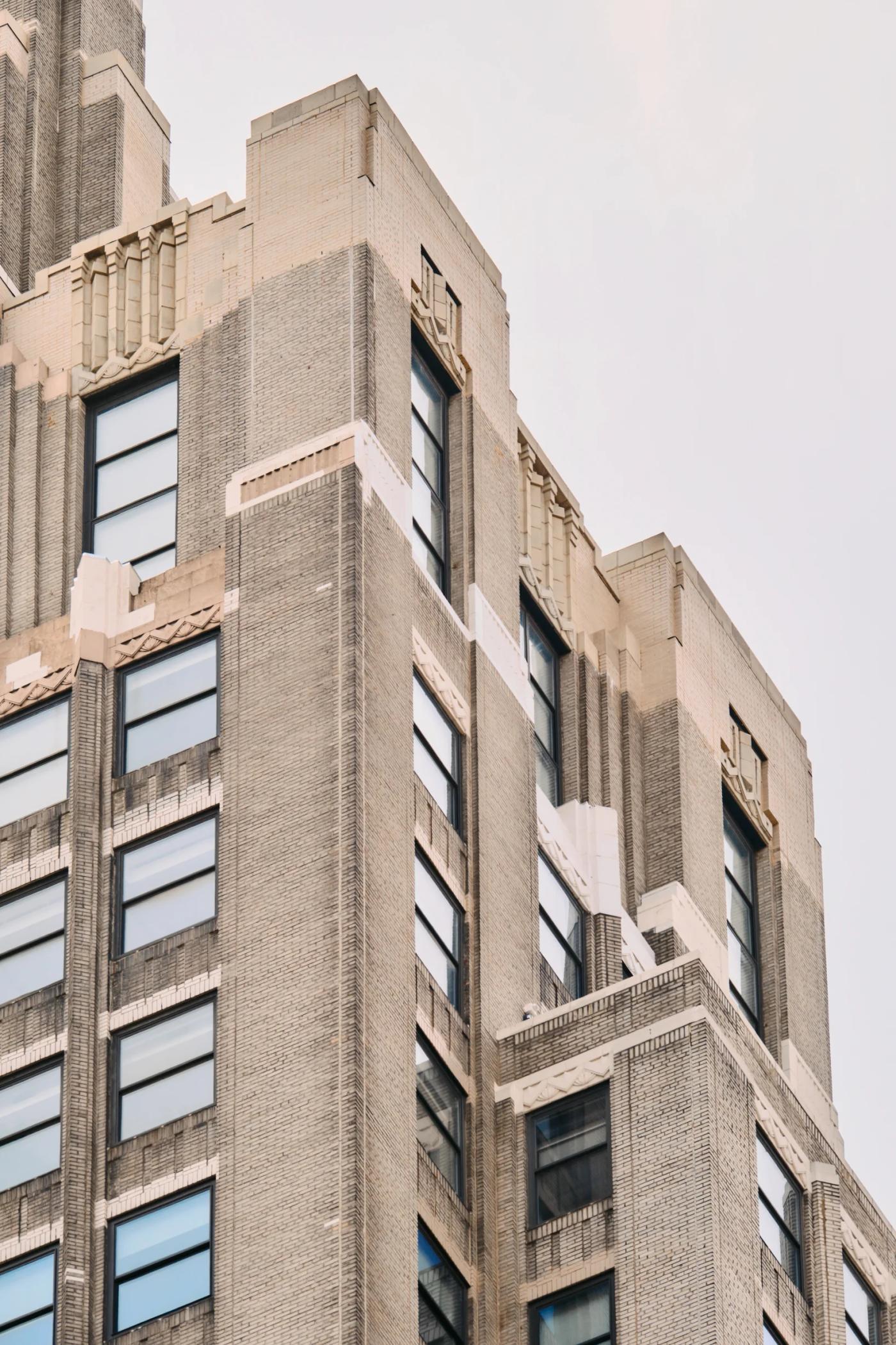 A tall, gray brick building with multiple levels and windows stands against a pale blue sky, captured from a low angle emphasizing its height and gran