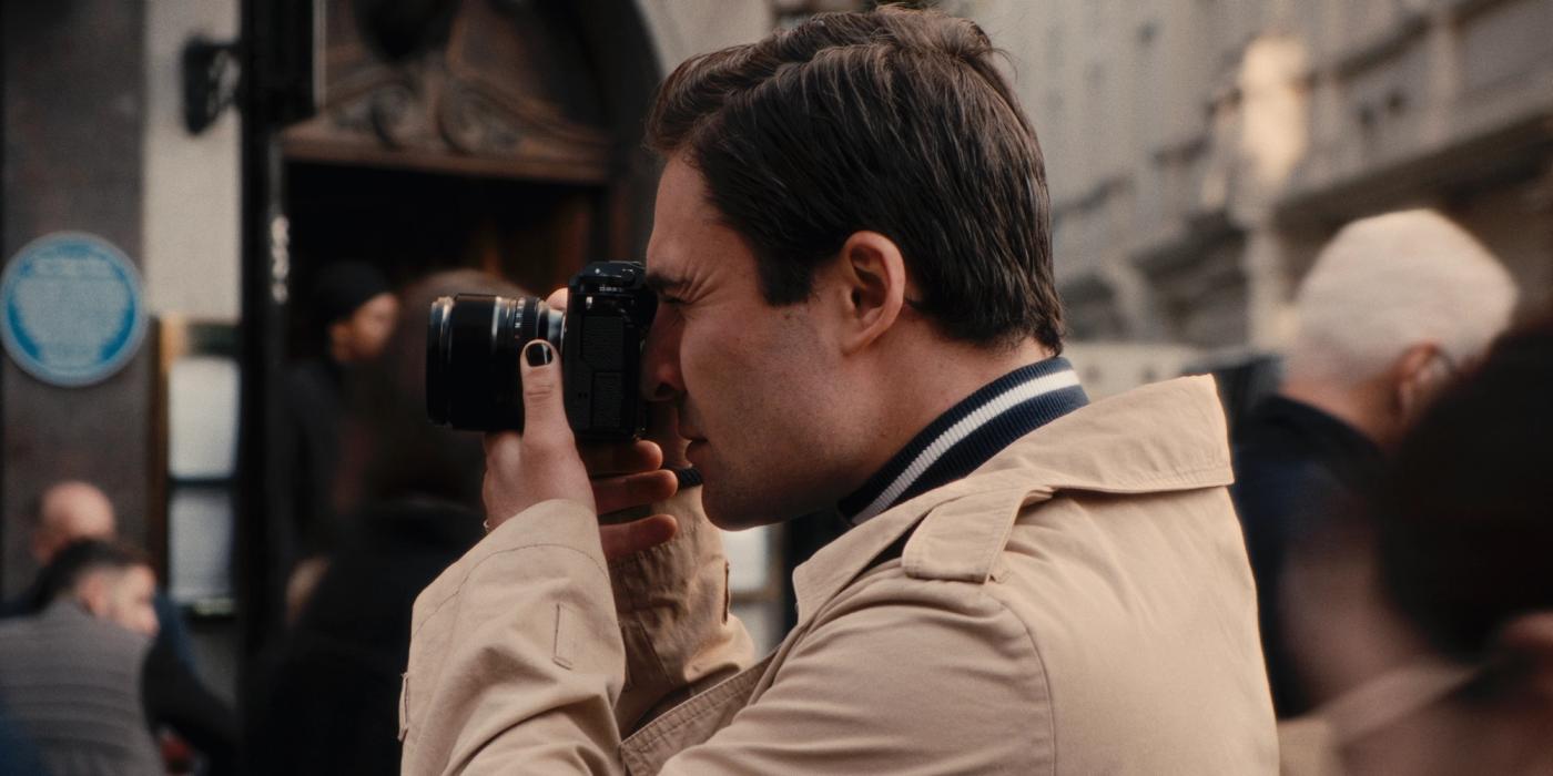 A man in a beige coat and black camera is taking a photo on a busy street, surrounded by people and buildings.