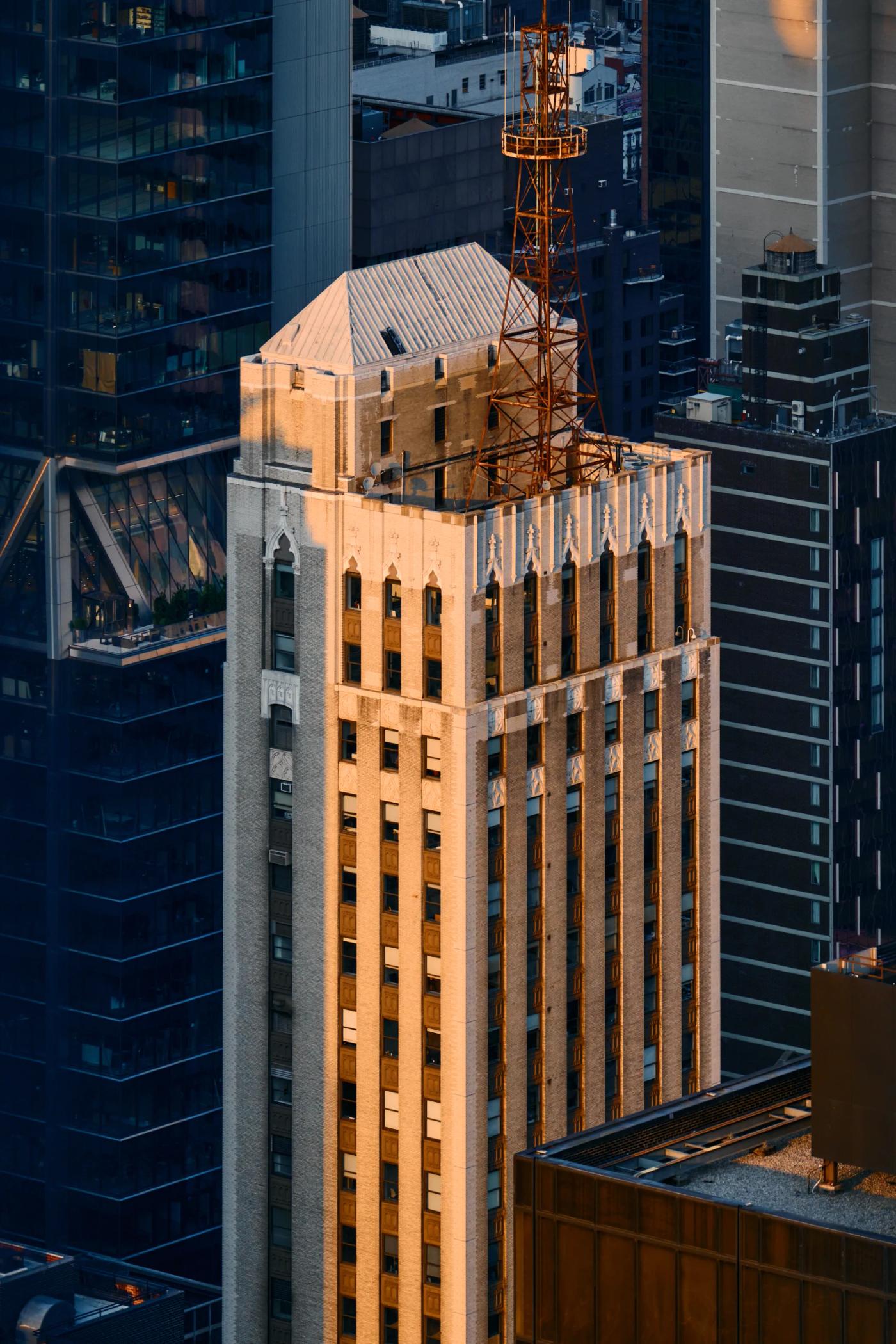 A tall, beige building with a white roof and orange crane stands in a bustling cityscape, surrounded by other buildings and a clear blue sky.