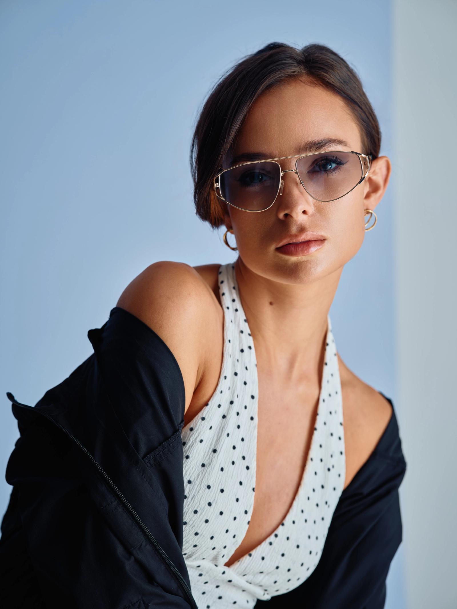 A young woman in a polka dot dress and sunglasses poses for a portrait against a blue background.