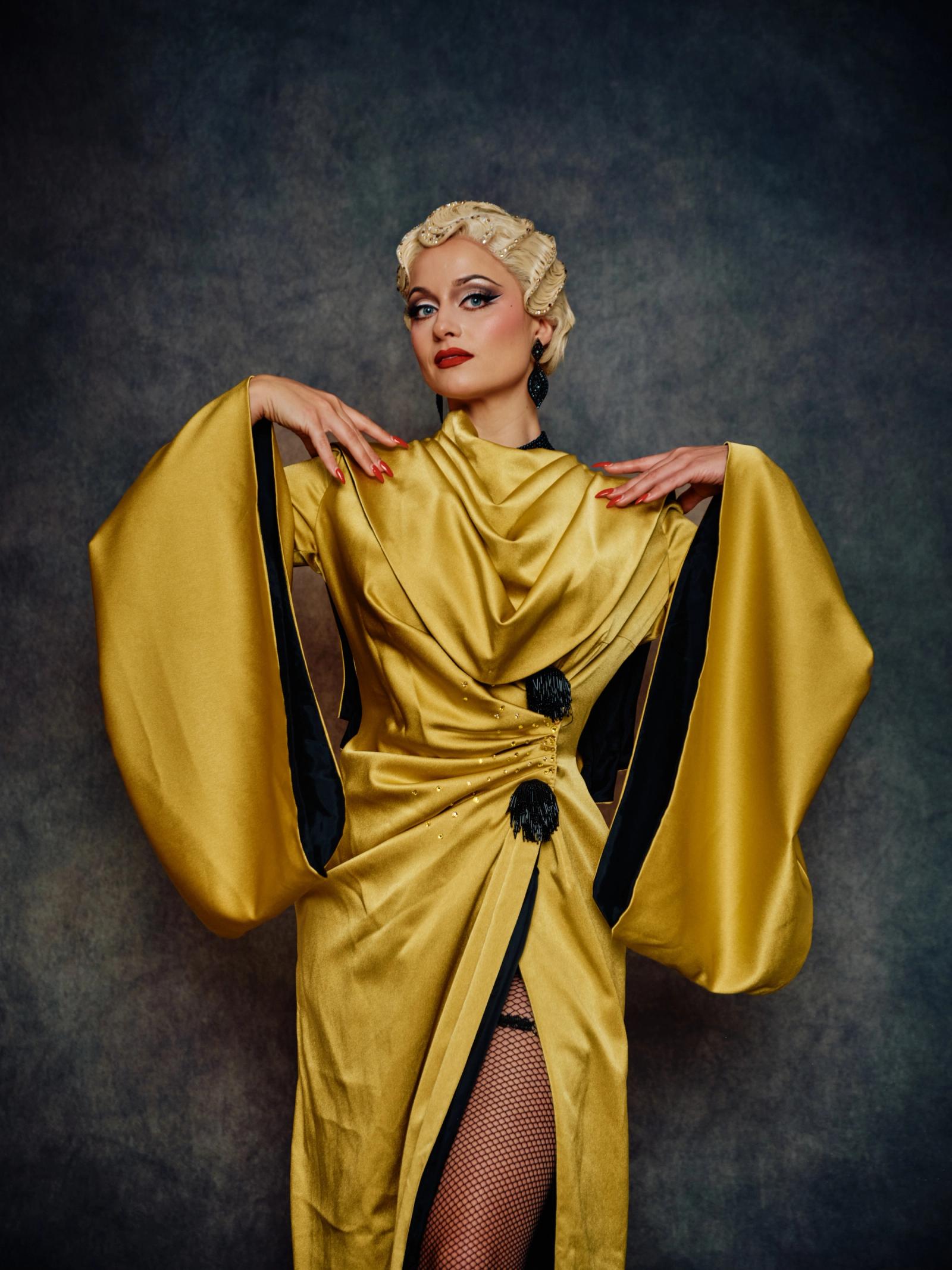 A woman in a golden, flowing dress with black accents and a black belt stands against a dark gray background, her blonde hair styled in a bob, and her