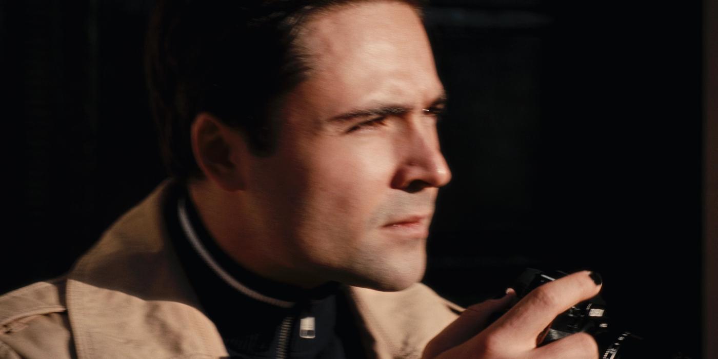 Actor Robert De Niro is captured in a moment of contemplation, dressed in a beige jacket and holding a black camera. His gaze is directed off to the s