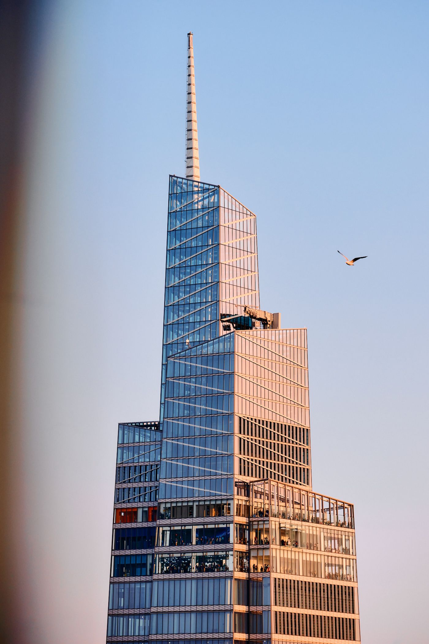 A skyscraper with a blue glass facade and a pointed spire is captured in a low-angle shot, showcasing its height and grandeur. A bird is seen flying i