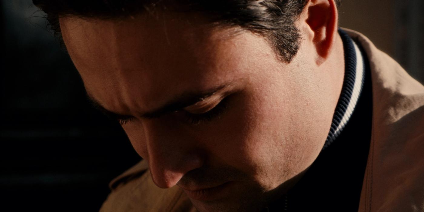 Man in beige jacket, dark hair, looking down, in dimly lit room.