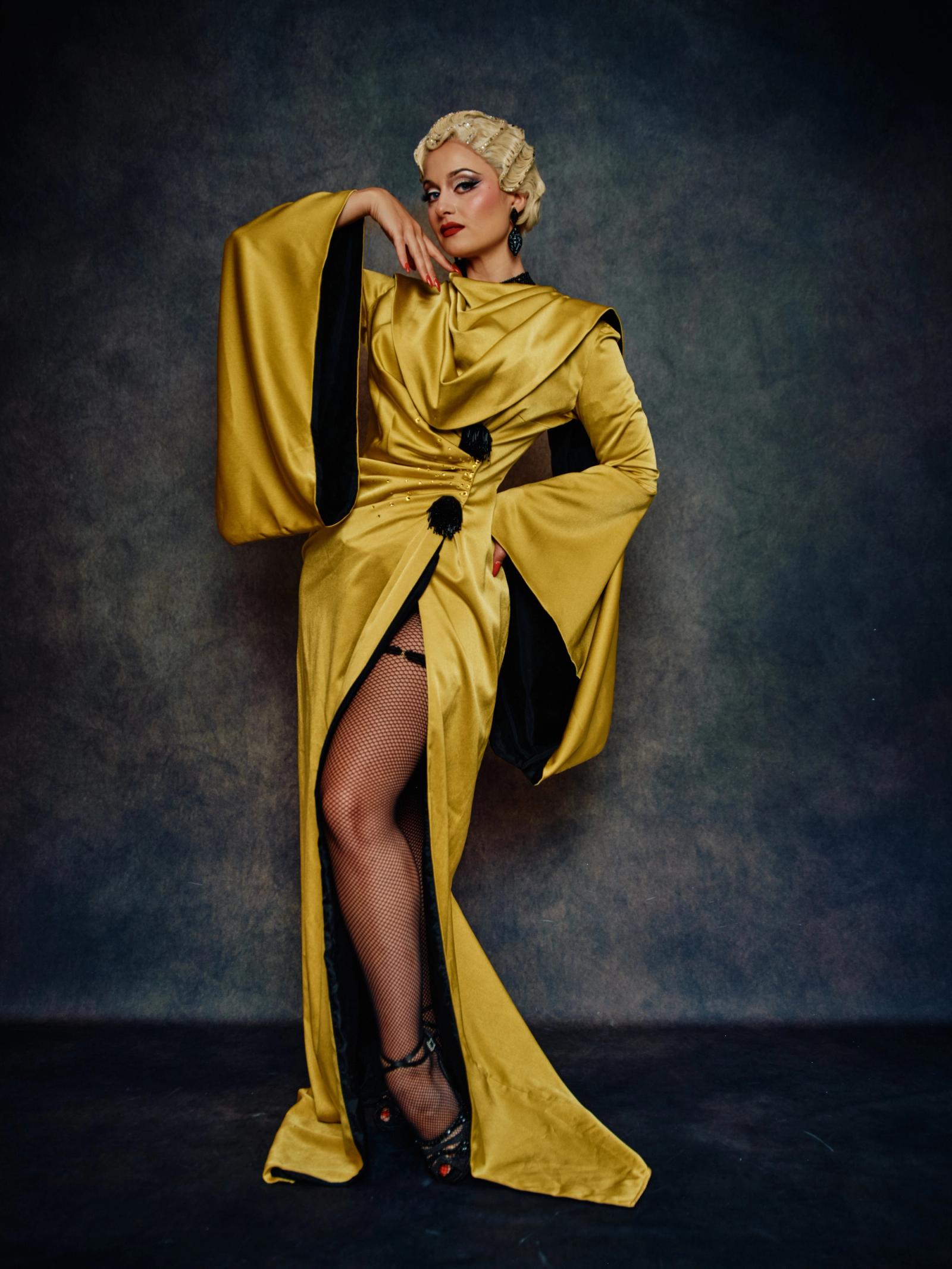 A woman in a golden, black, and gold dress with a black belt and black heels poses elegantly against a dark blue background.