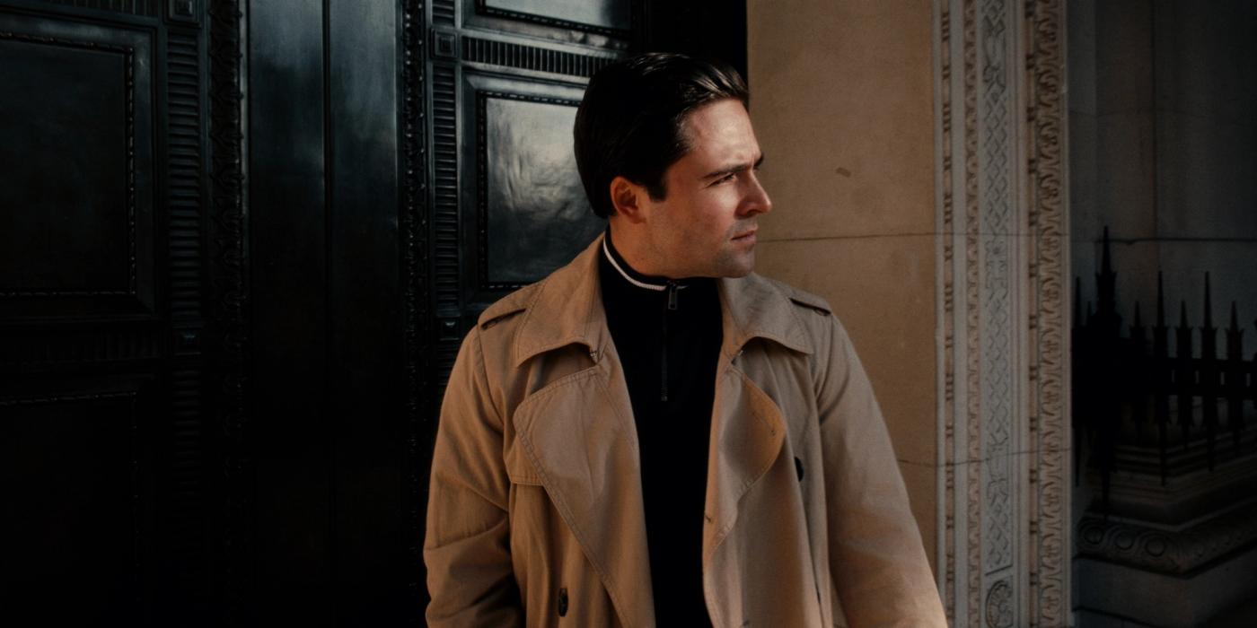 Actor Patrick Stewart stands in front of a black door, wearing a beige trench coat and black shirt. His gaze is directed off to the side, and he appea