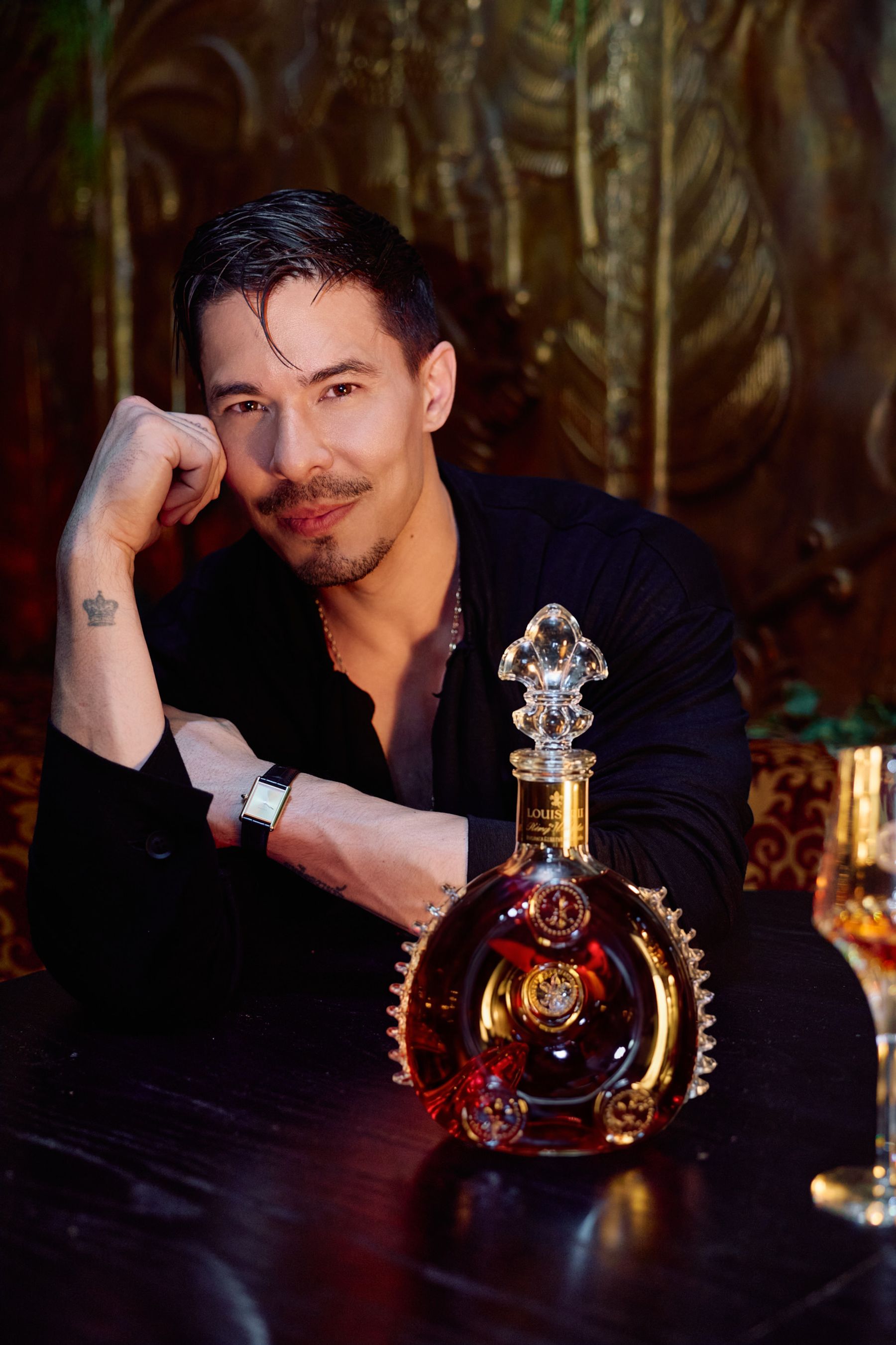 Man with tattoos, wearing a black shirt, sits at a table with a bottle of liquor, a wine glass, and a watch. Louis XIII Cognac