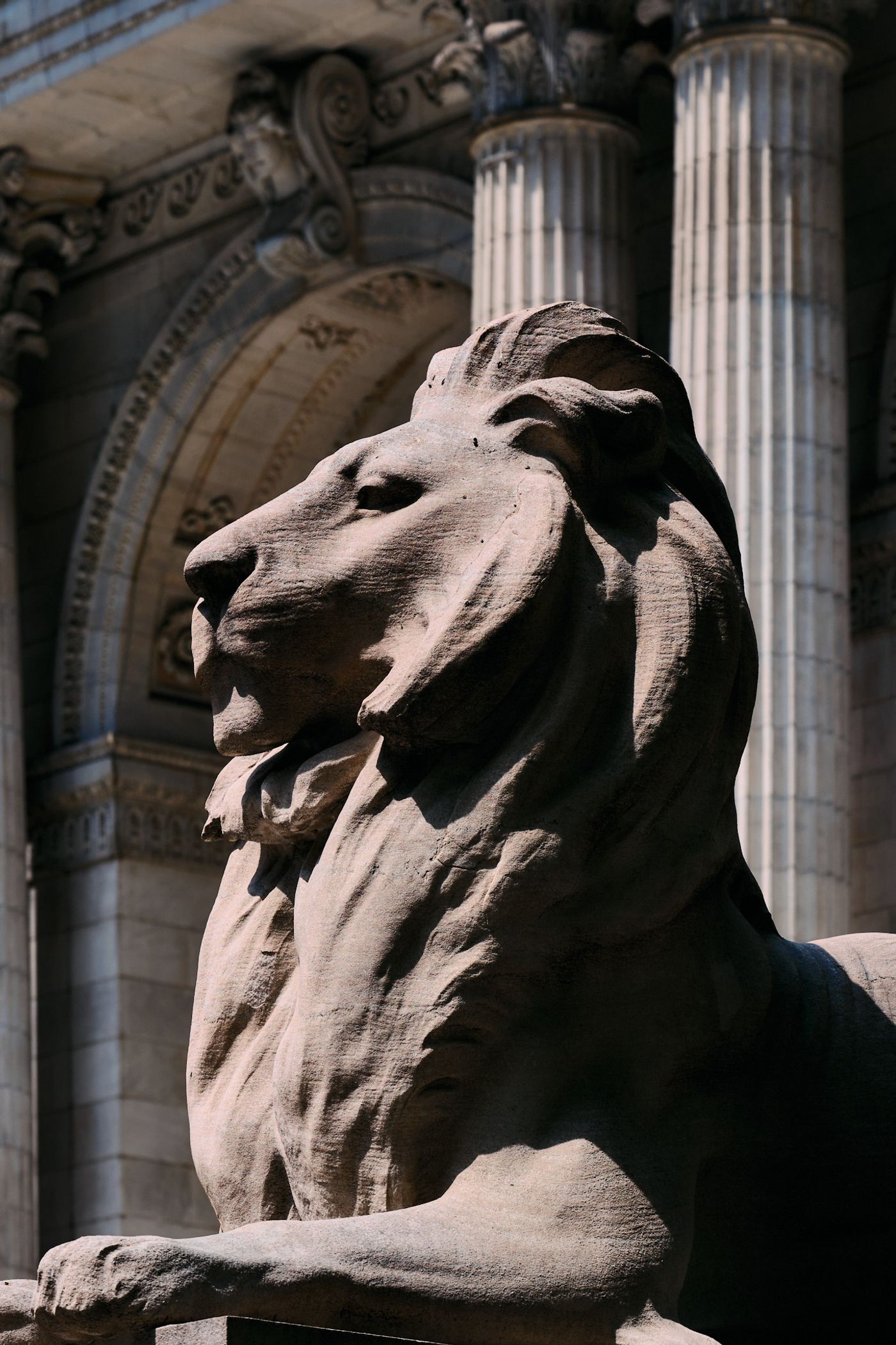 A majestic stone lion statue stands tall in front of a grand, ornate building with columns and intricate carvings. The lion's head is turned to the le