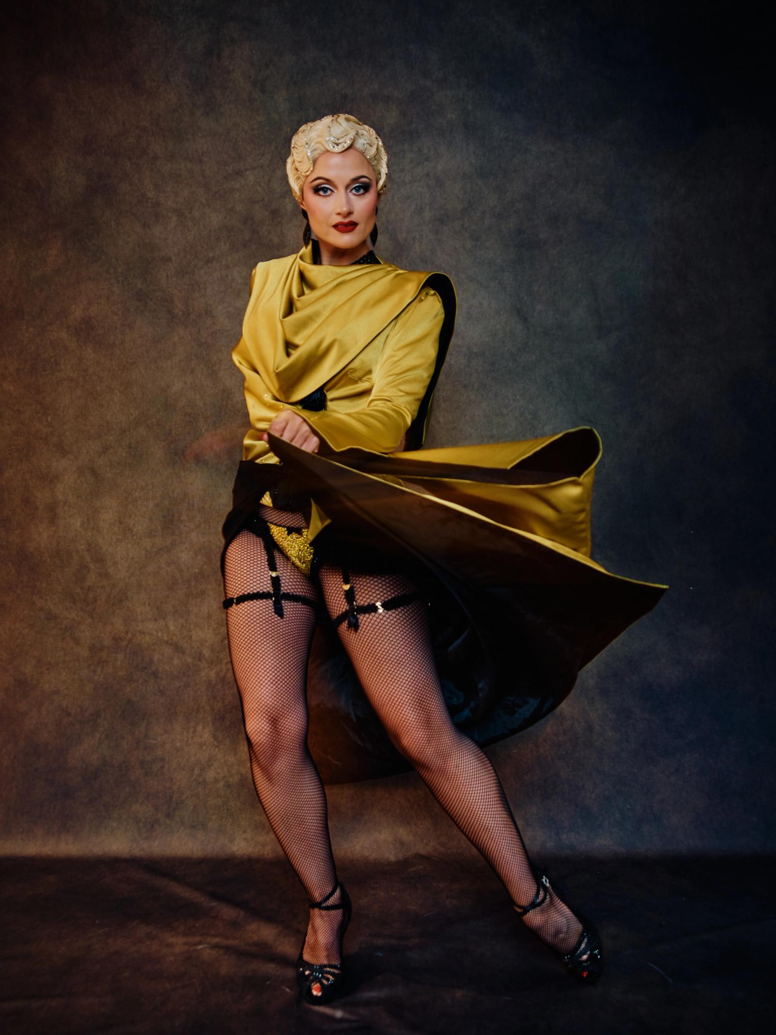 A blonde woman with blonde hair, wearing a yellow top, black skirt, and black high heels, poses confidently with her left hand on her hip and her righ