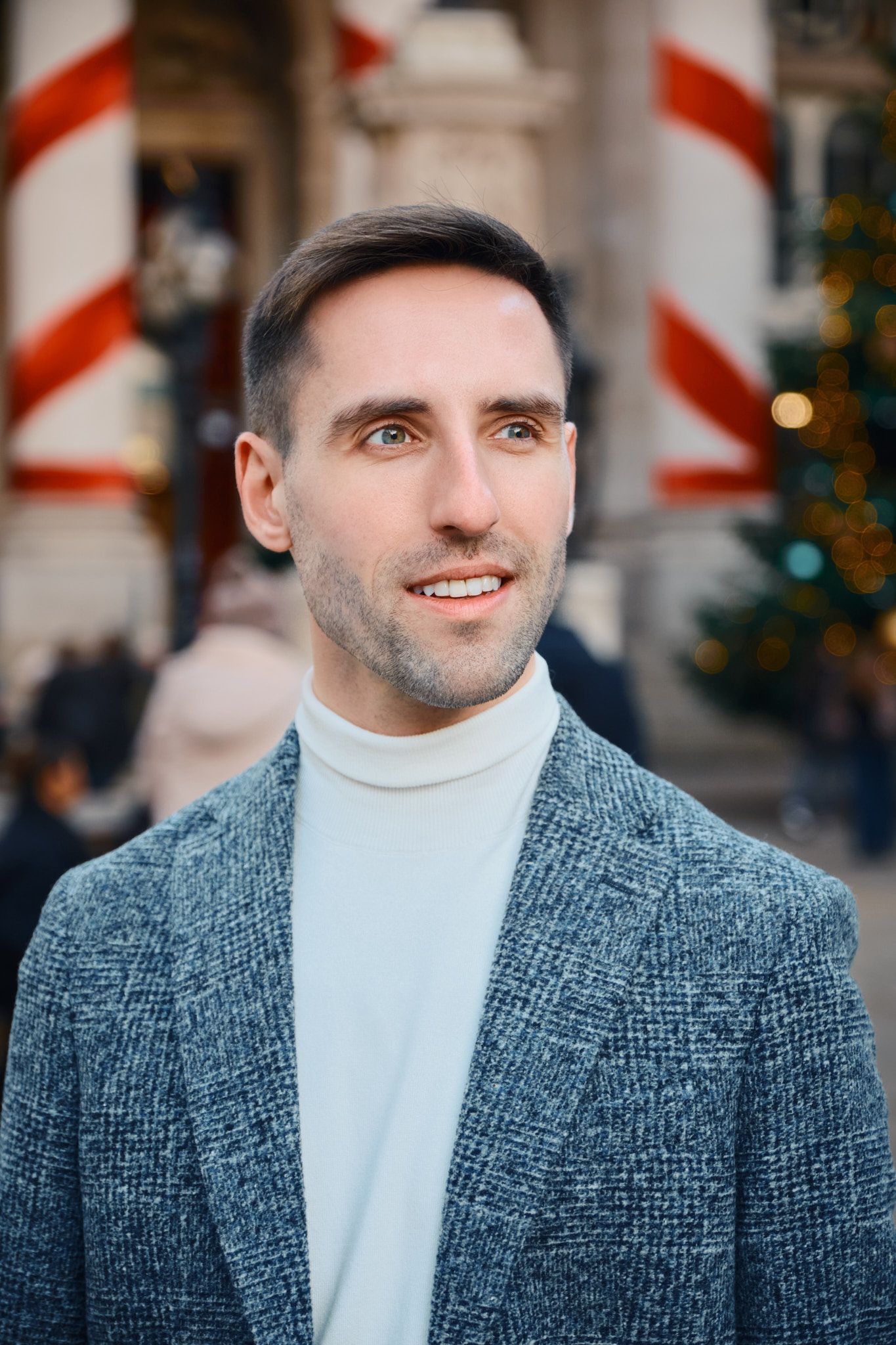 A man in a gray blazer and white turtleneck stands in front of a grand building with columns and a Christmas tree, gazing at the camera.