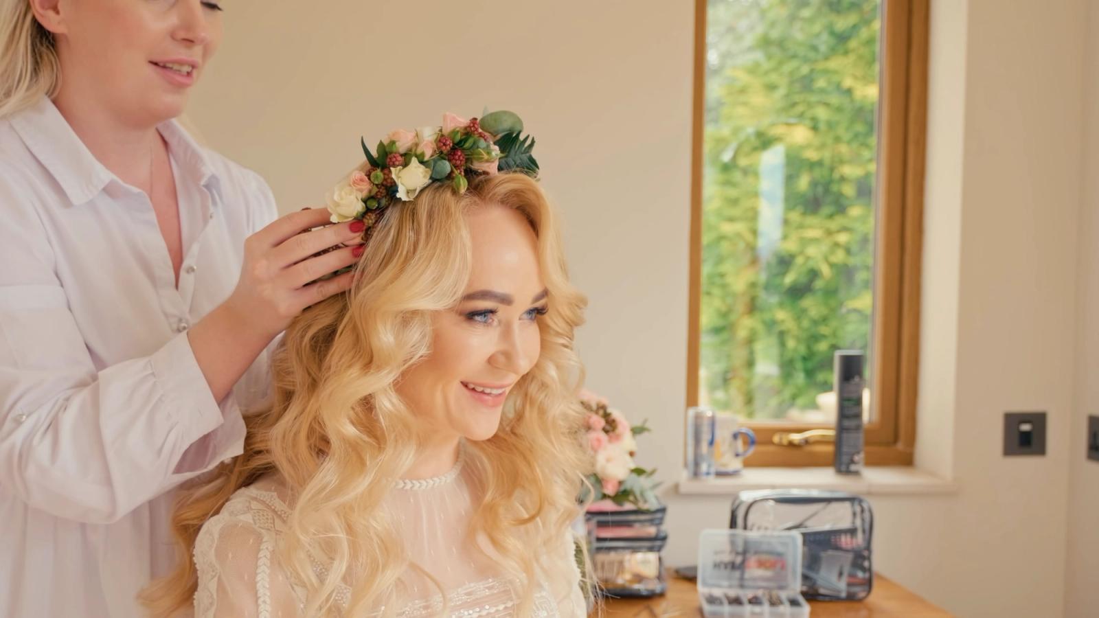 A woman with long blonde hair is getting her hair styled by a woman in a white shirt. They are in a room with a window, a mirror, and a counter with a