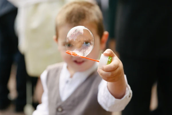 Close-up candid portrait of a young boy in a waistcoat holding a soap bubble wand with a large bubble in front of his face at the wedding. Playful documentary moment.