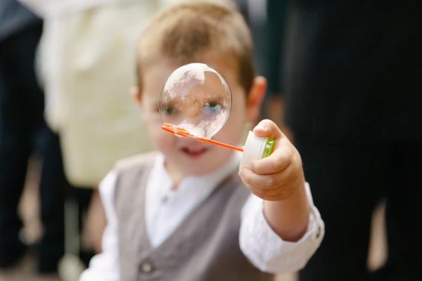Close-up candid portrait of a young boy in a waistcoat holding a soap bubble wand with a large bubble in front of his face at the wedding. Playful documentary moment.