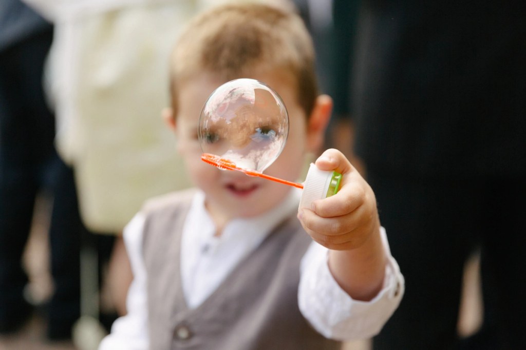 Close-up candid portrait of a young boy in a waistcoat holding a soap bubble wand with a large bubble in front of his face at the wedding. Playful documentary moment.