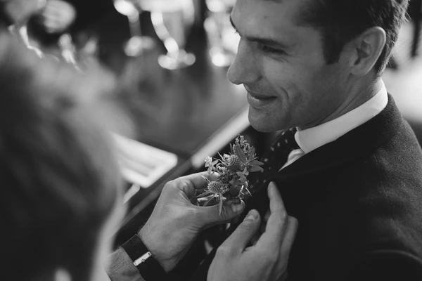 Groom smiling at the bouquet during the ceremony — white shirt and tie, guests blurred behind