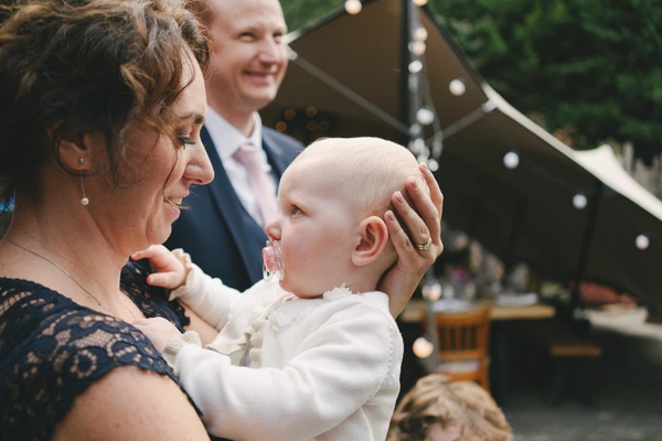 Mother tenderly holds her little daughter in the wedding marquee, groom watching with a smile