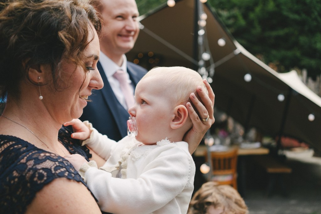 Mother tenderly holds her little daughter in the wedding marquee, groom watching with a smile