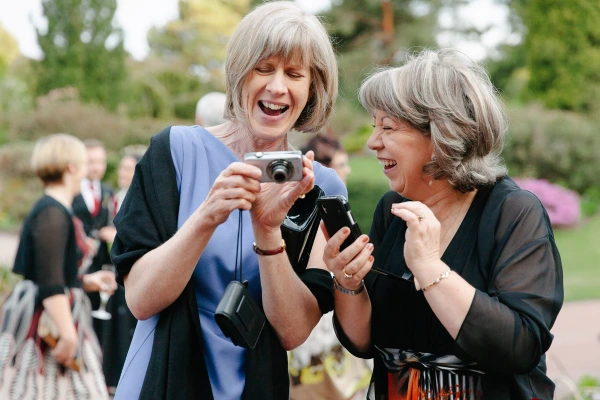Two women laugh together while looking at photos on a compact camera at the wedding drinks reception outdoors in the garden. Candid documentary wedding moment.