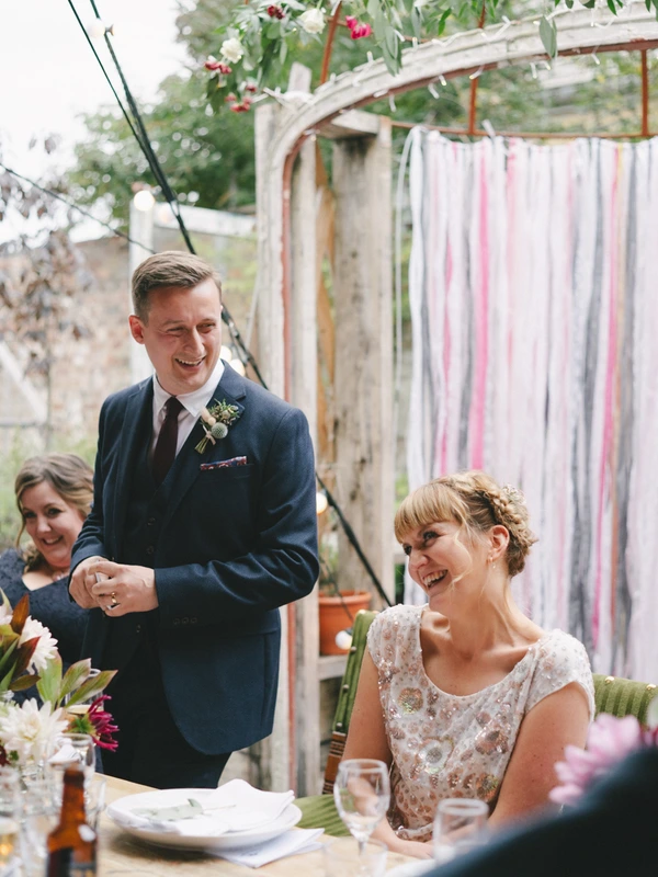 Groom stands smiling by the table, bride seated looking up at him — garden pavilion, reception