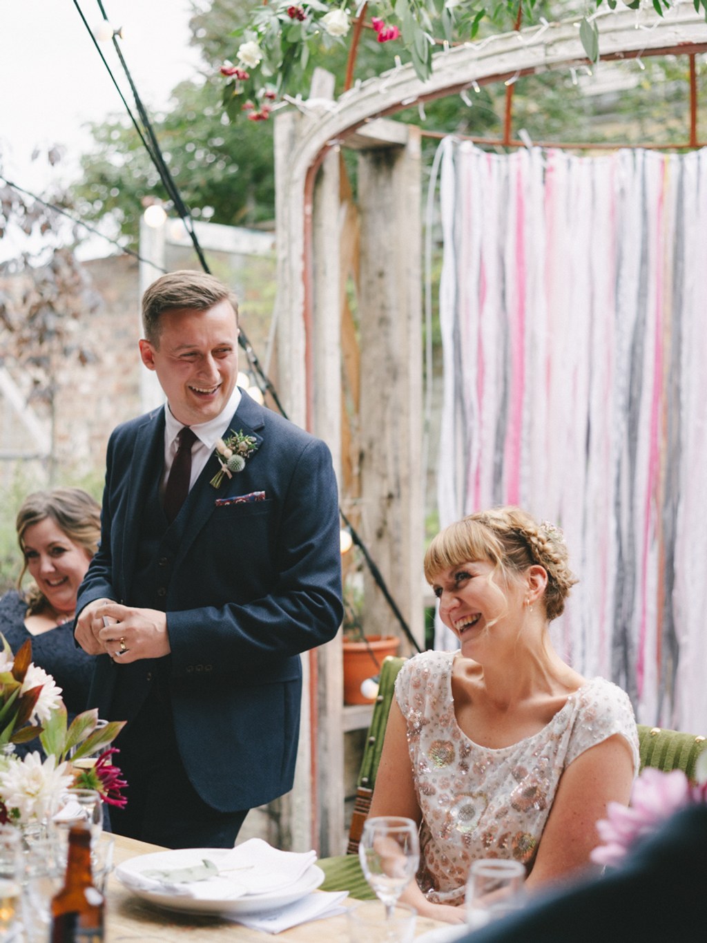 Groom stands smiling by the table, bride seated looking up at him — garden pavilion, reception
