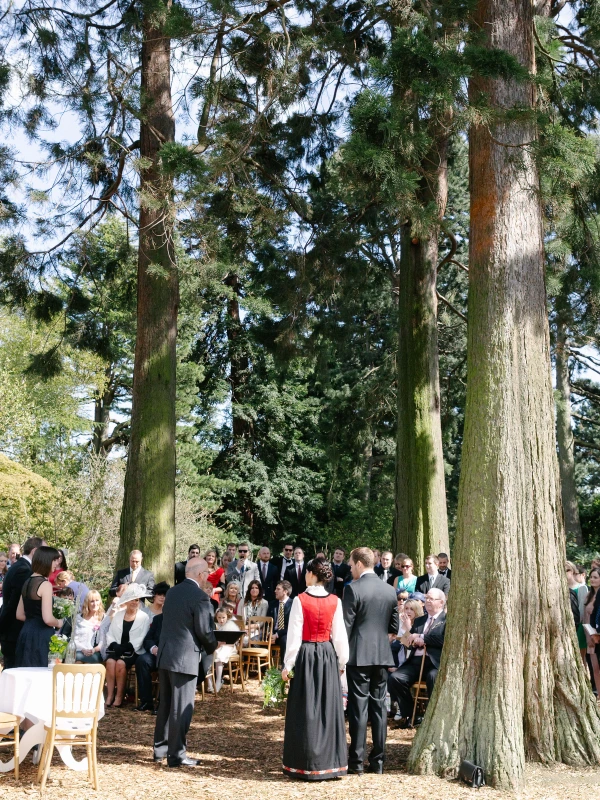 Wide colour shot of the outdoor wedding ceremony among giant redwood trees at the Royal Botanic Garden Edinburgh, bride in red Norwegian bunad standing with the groom. Documentary overview.