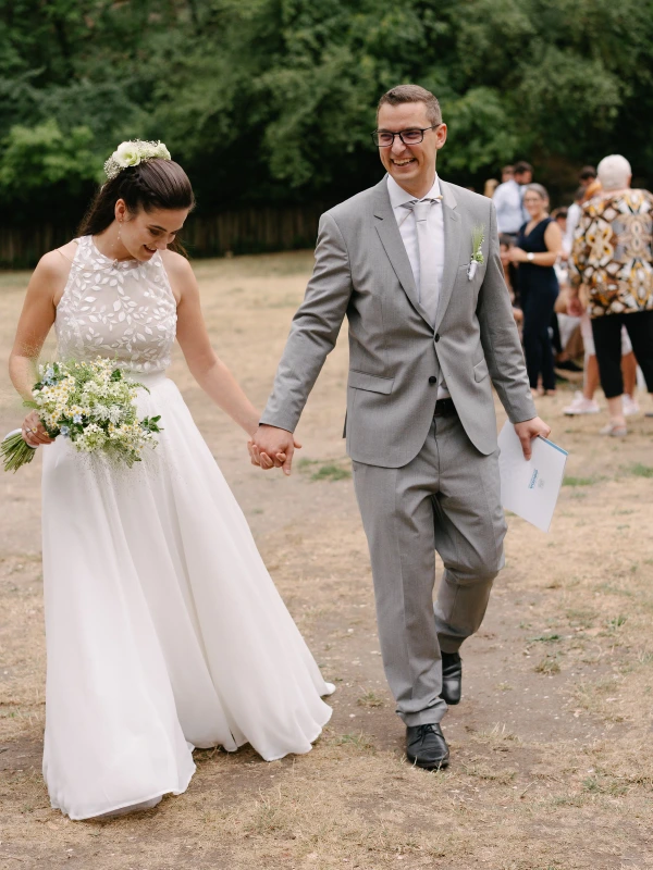 Bride holding white daisy bouquet walking with groom after wedding ceremony outdoor garden venue.