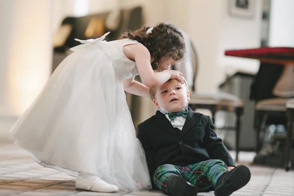 Bride stoops toward a little boy in black suit and tartan trousers — warm natural light