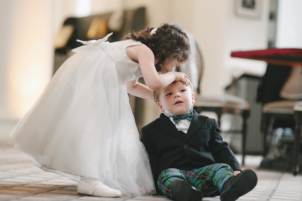Bride stoops toward a little boy in black suit and tartan trousers — warm natural light