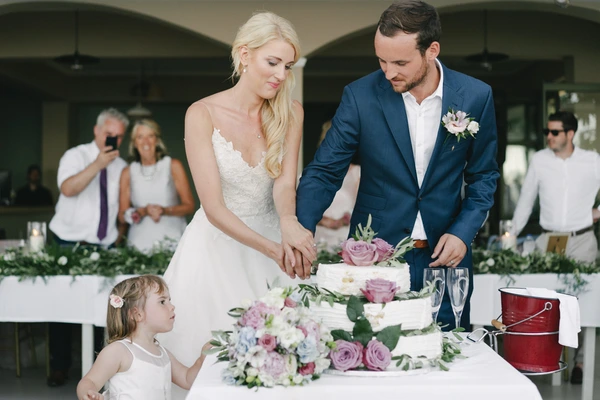 Bride and groom cut the cake — white lace, blue suit, little girl watching, pink and white tiered cake