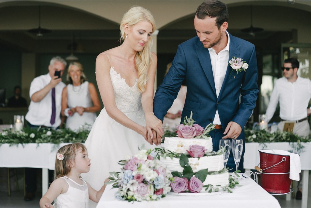 Bride and groom cut the cake — white lace, blue suit, little girl watching, pink and white tiered cake